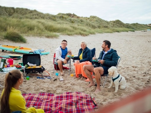A family and their dog sat beside a picnic blanket on beach, Beadnell, Northumberland