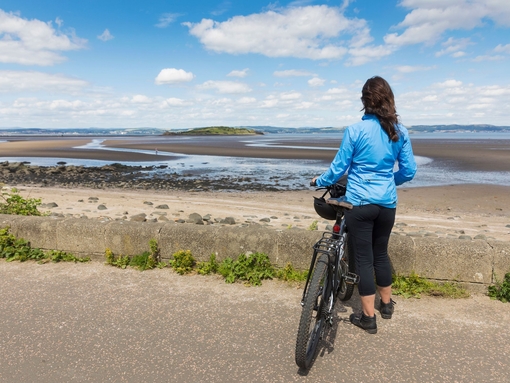 A female cyclist holding a bike and looking out over a beach. 