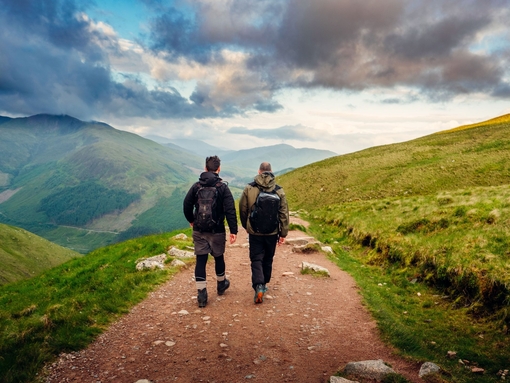 Two male hikers trekking along a pebbled path, cloudy skies, green rolling mountain vista.