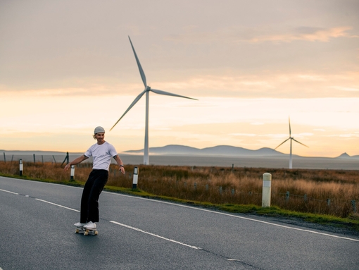A male skateboarder enjoys the flat road surface to skate on in the Scottish Highlands, with wind turbines behind him
