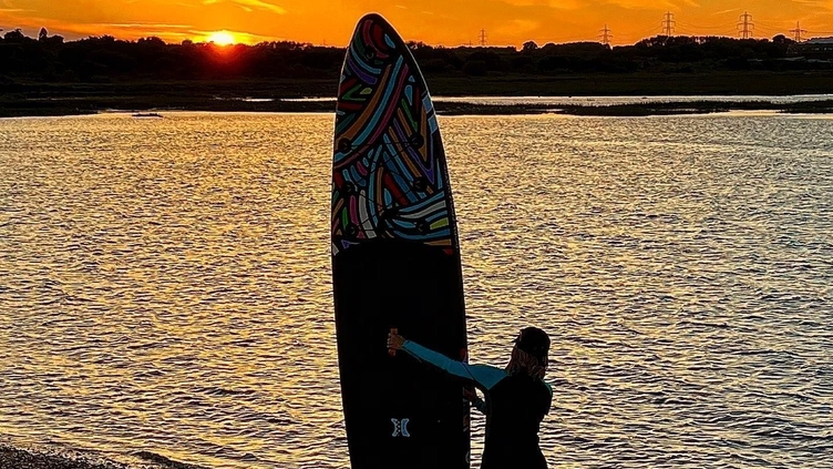 Person holding a paddle board on the shoreline at sunset