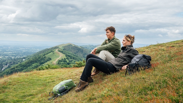 Two women sit on a hill looking toward a view with green hills beyond