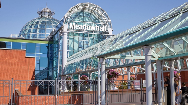 People walking under a glass roofed tunnel into the entrance of a shopping centre.