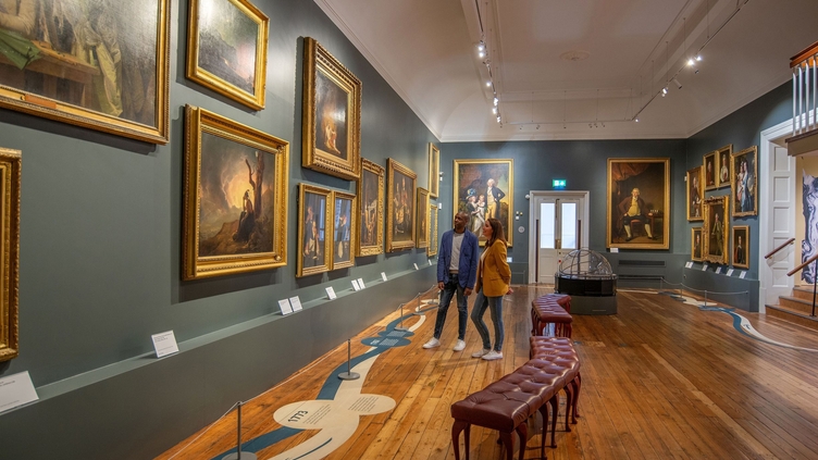 A man and a woman stand looking at paintings in a gallery