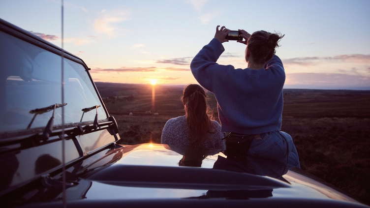 Women sitting on bonnet of 4x4 parked on road at sunset