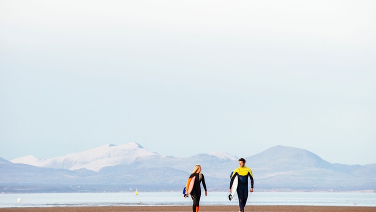 Man and woman, in wetsuits, carrying surfboards on beach