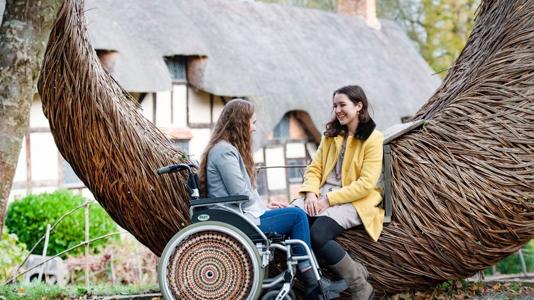 Wheelchair user and friend sat beside a wooden sculpture