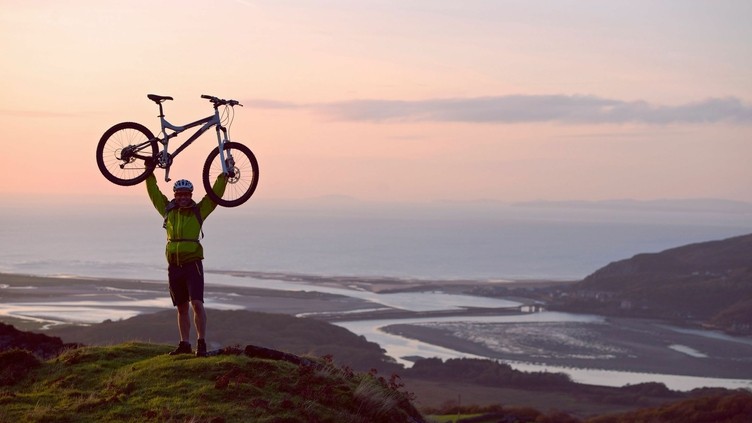 Ein Mann, der sein Mountainbike feierlich über den Kopf hält, auf dem Gipfel eines Hügels mit Blick auf die Küste und das Meer unter ihm