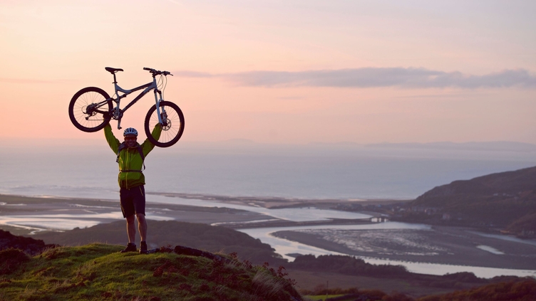 Man holding his mountain bike above his head in celebration at the top of a hill with the coast and sea below him
