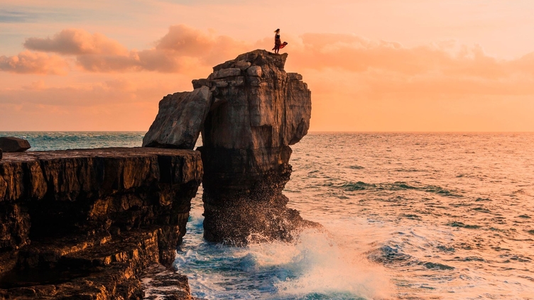 Woman standing on a rock stack looking out to sea at sunset