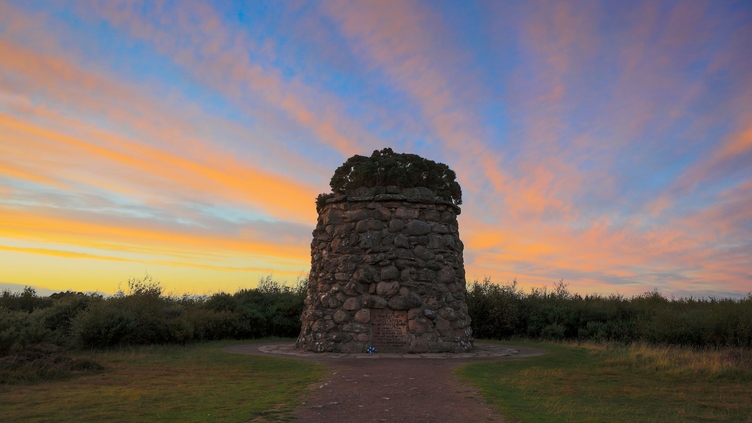 A small stone tower with a plaque on it surrounded by grass and moor under a vibrant blue and orange sky at sunset.