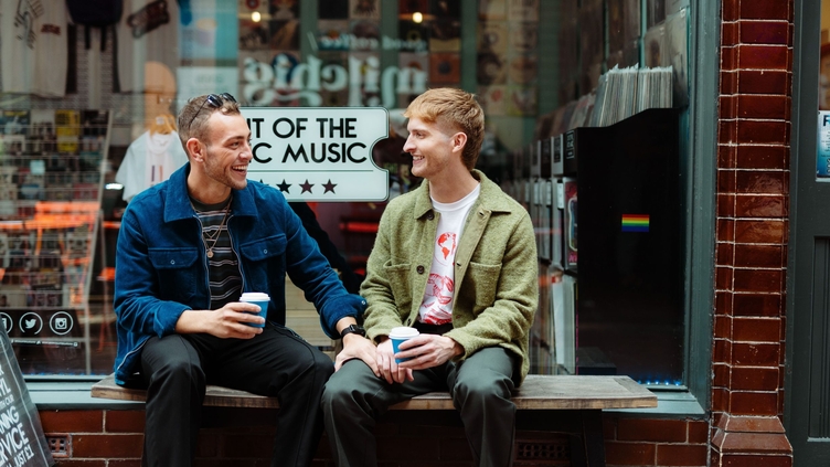 Two men sit with take away drinks in front of a shop