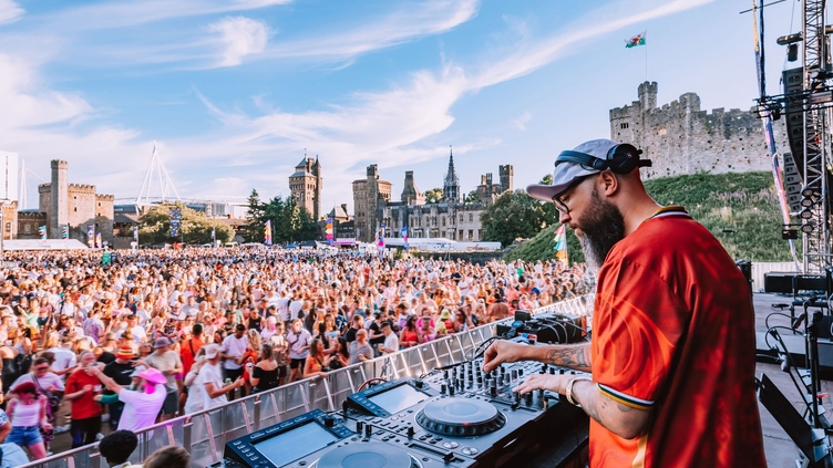 A DJ using music decks on a stage overlooking a crowd in the grounds of a castle