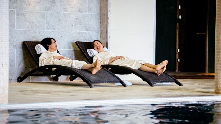 Two women, in white robes, lying down beside a swimming pool