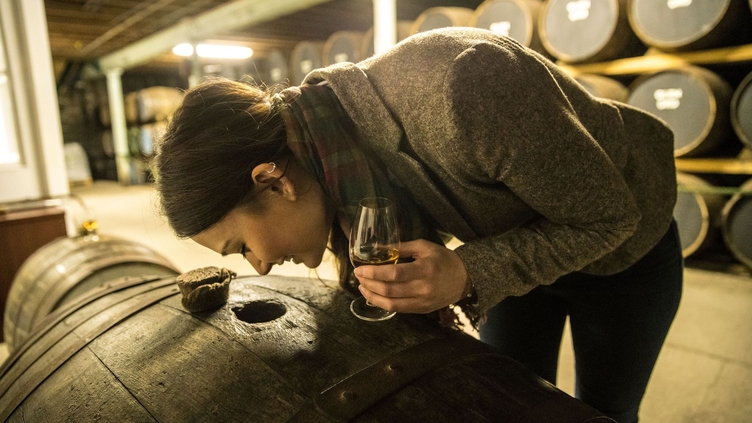 Young woman smelling whisky through a hole in an oak barrel