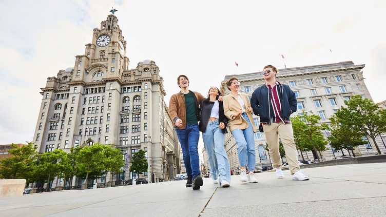 Four friends exploring a local city walking along waterfront promenade, with historic buildings in the background