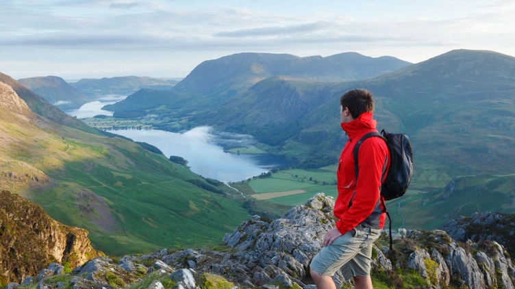 A man wearing a rucksack stands on a mountain fell overlooking hills and a lake