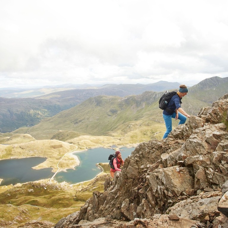Two females hiking together up a large mountain range, with a lake below