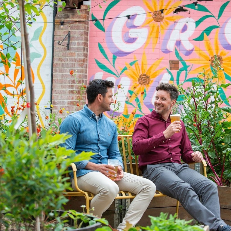 Two men sat on a bench in a garden with a drink