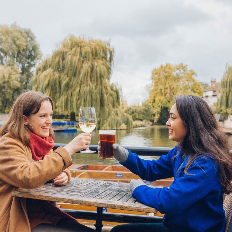 Two women toasting with drinks at a pub beside the river