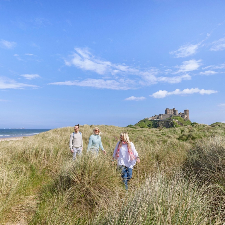 A group of people walk among tall grasses in dunes with a castle in the background