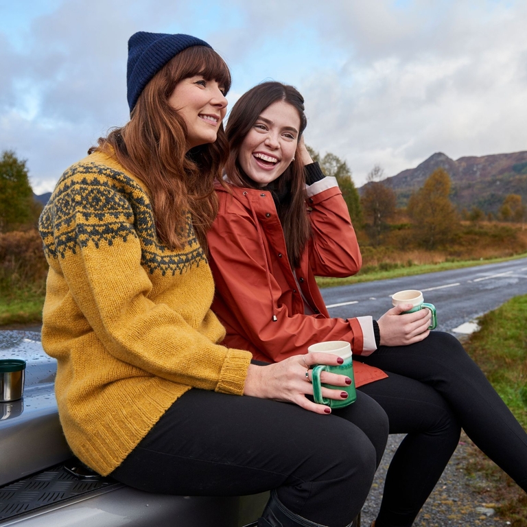 Zwei Frauen machen an einer Landstraße in den schottischen Highlands Zwischenpause und trinken Kaffee aus einer Thermoskanne.