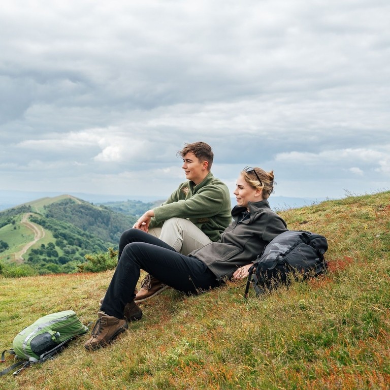 Two women sit on a hill looking toward a view with green hills beyond