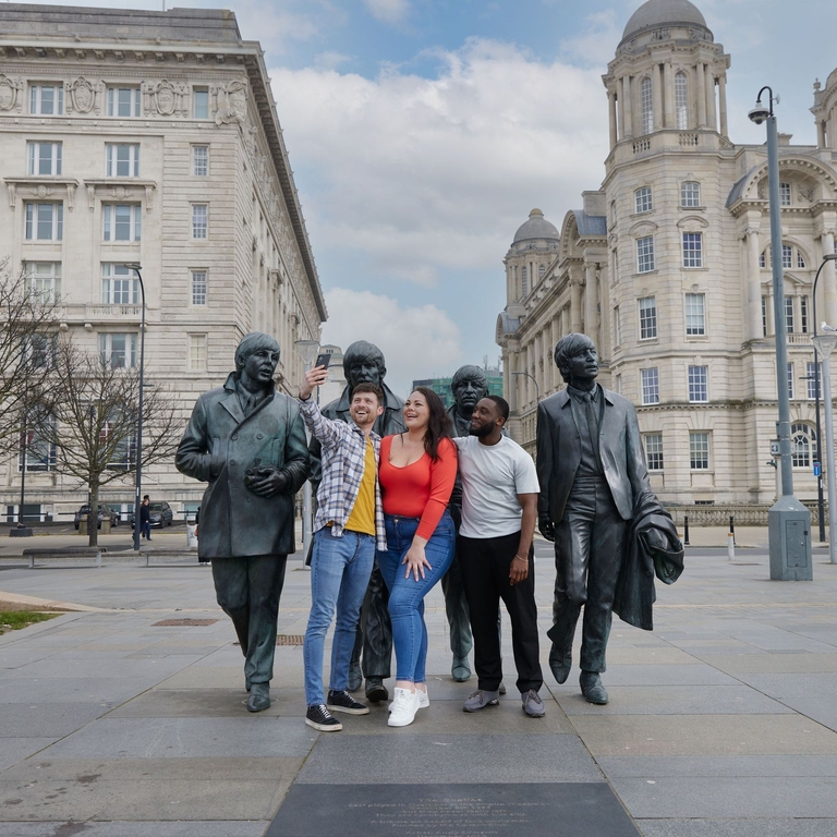 A group of friends, two men and a woman take a selfie in front of a statue in a city centre.