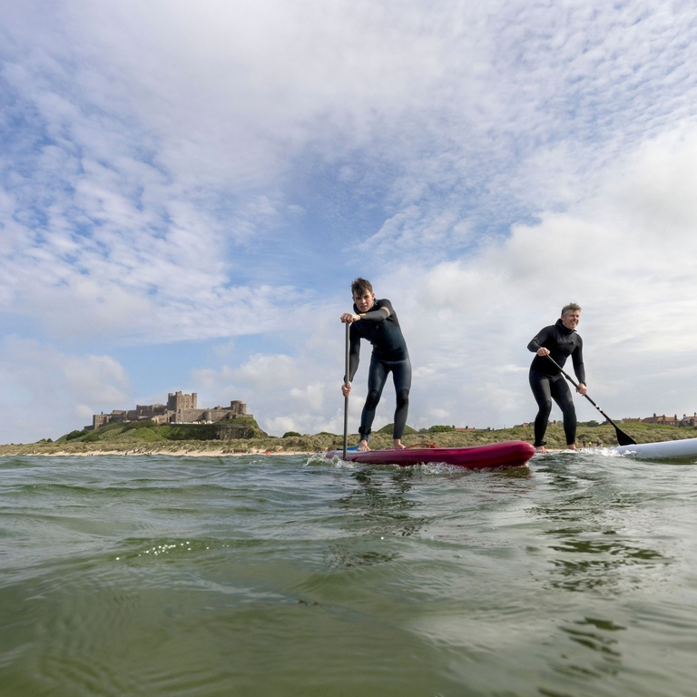Two men on are paddleboarding in the sea with a heritage castle in the background.