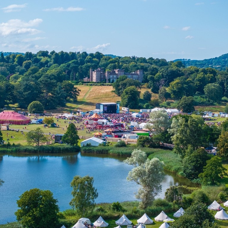 A large outdoor concert with a large screen with crowds and many tents and fairground rides around a large lake.
