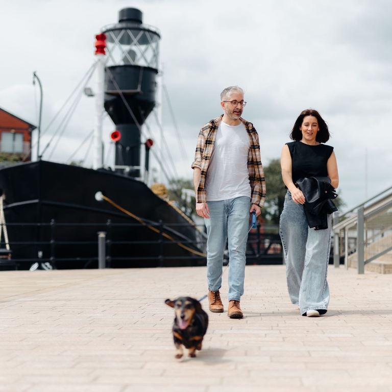 A man and a woman walk their dog in front of a large boat