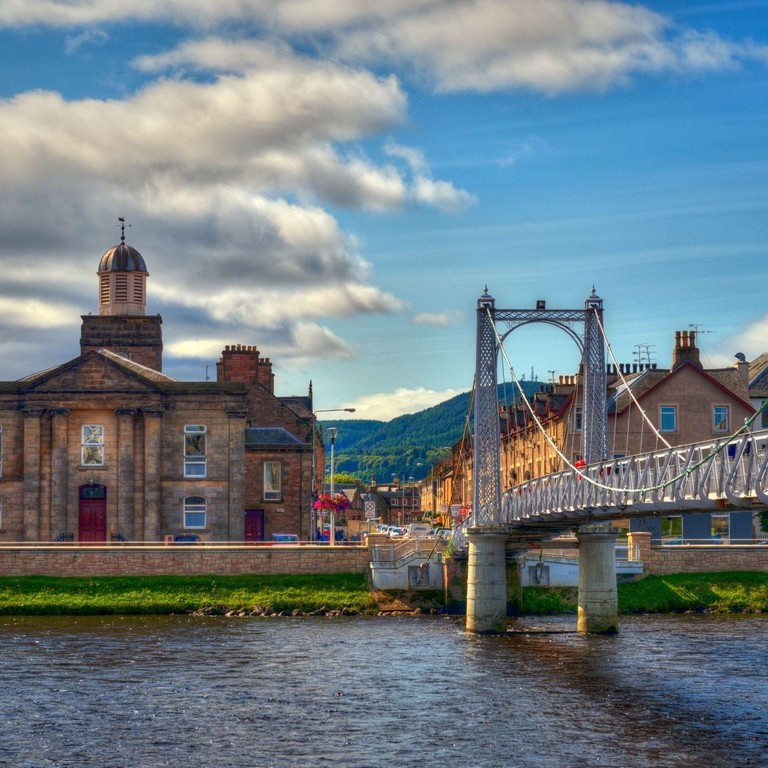 A suspension bridge with people walking across spanning a wide river with houses on the other side