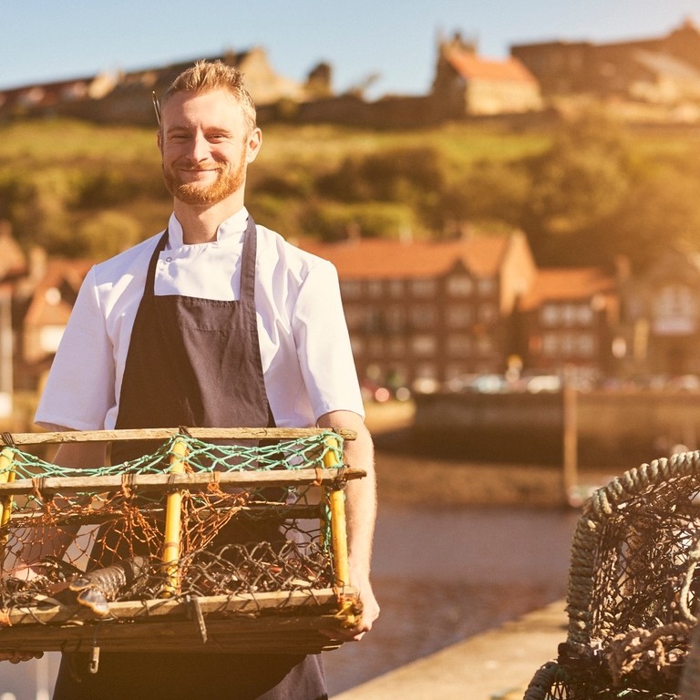 Chef wearing apron on pier holding lobster trap with lobster