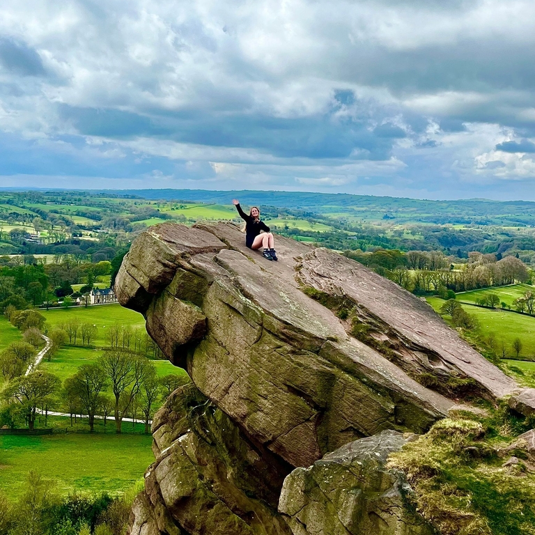 A seated woman waving from a hanging stone, overlooking a country panorama.