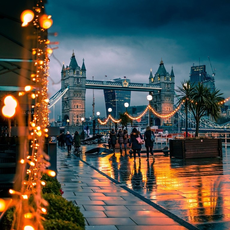 Fußgänger, die an einem verregneten Abend an Lichterketten vorbeigehen, im Hintergrund die Tower Bridge, London