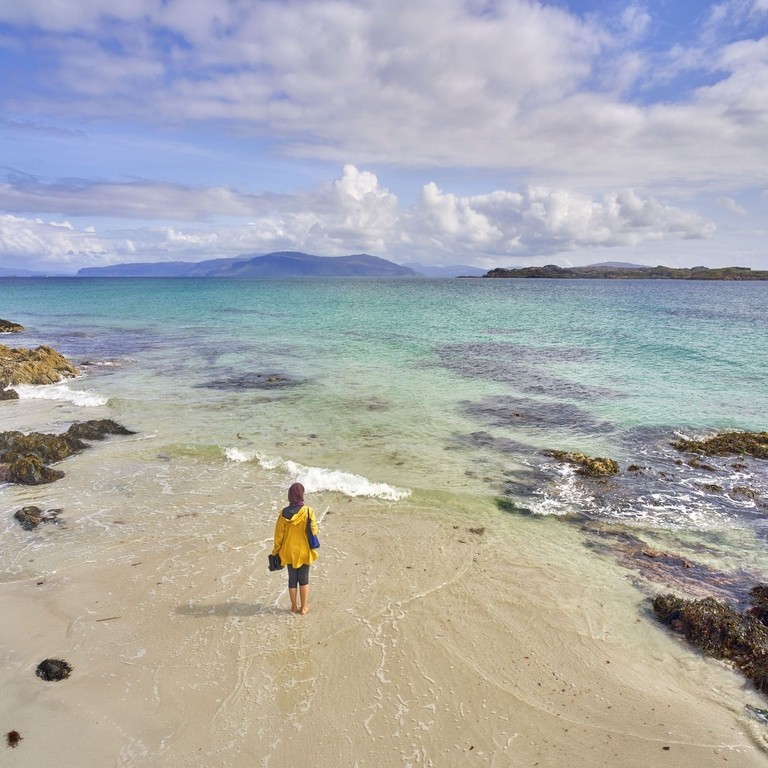 A high angle view of an independent young woman walking on the white sand with the crystal clear blue waters of Iona, Scotland