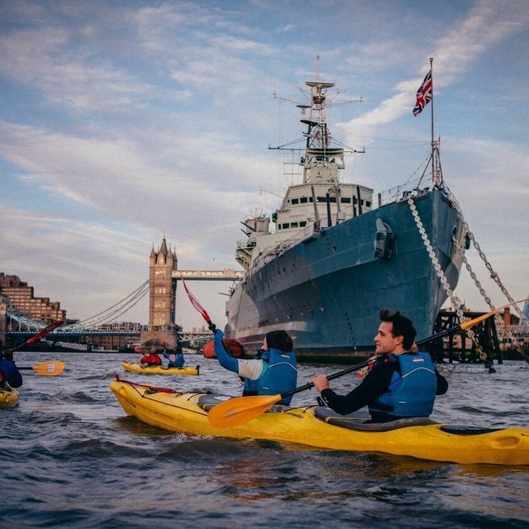Personnes en kayak sur la Tamise passant devant le HMS Belfast avec le Tower Bridge au loin