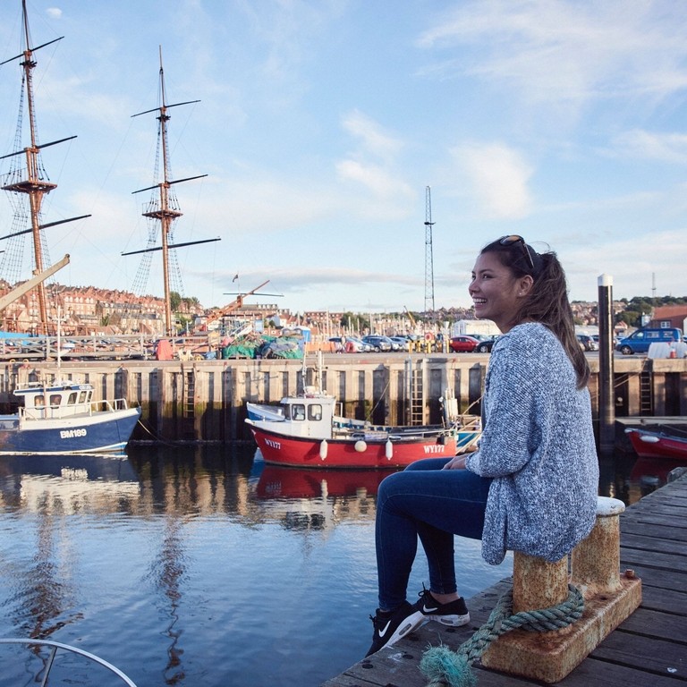 Smiling woman sitting on jetty on Whitby harbour looking at boats