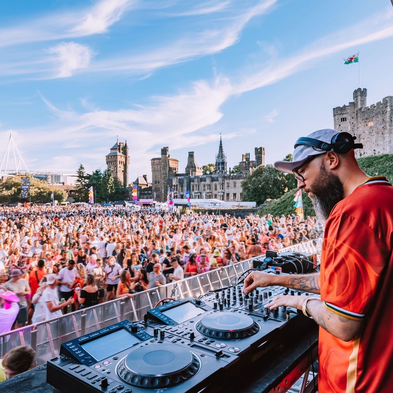 A DJ using music decks on a stage overlooking a crowd in the grounds of a castle