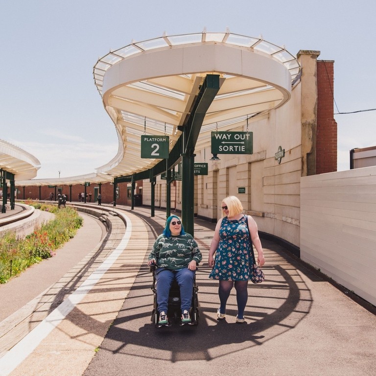 A woman walks with another woman who sits in a wheelchair at an old railway station