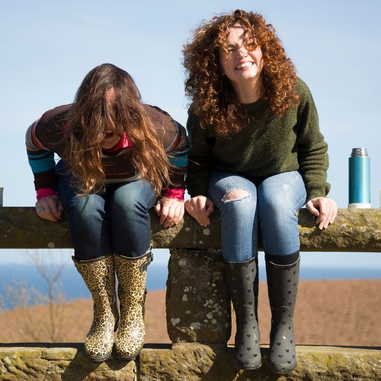 Two cheerful women holding mugs with hot drinks, laughing