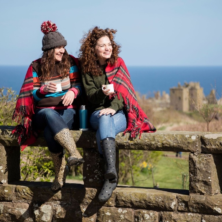 Women holding mugs with hot drinks, sitting on a stone wall