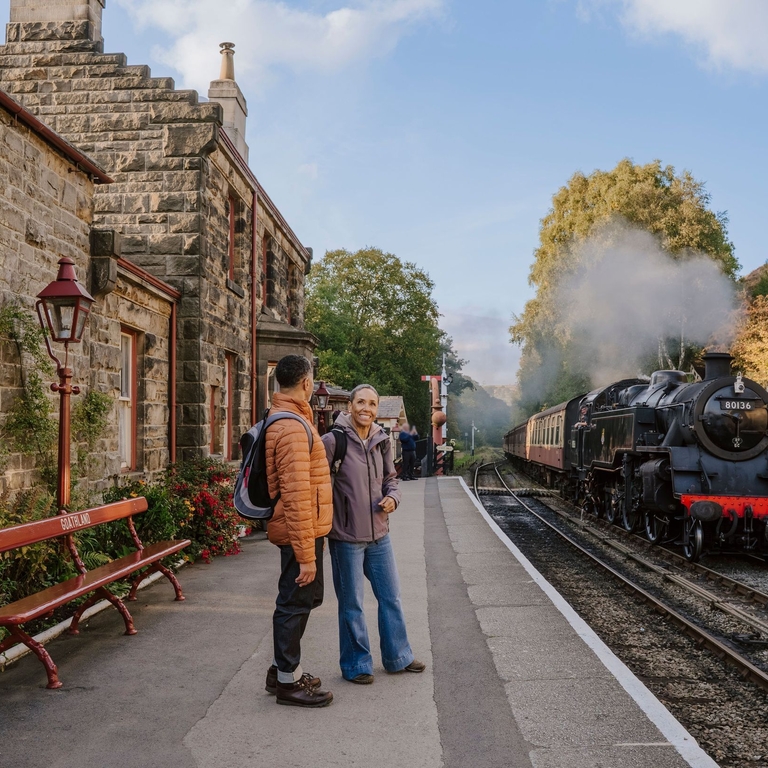 A man and woman standing on a train station platform as a train approaches in the background