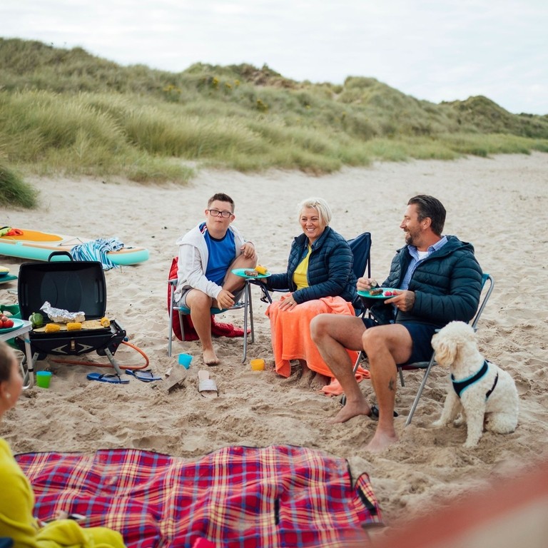 A family and their dog sat beside a picnic blanket on beach, Beadnell, Northumberland