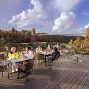 Friends enjoying tea and cake on a rooftop terrace, overlooking a river and suspension bridge.