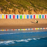 A surfer walking the beach in front of colourful beach huts