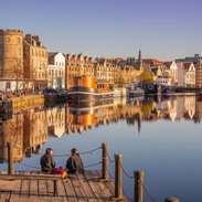 Two young women sat on the jetty of the waterfront in the evening with boats and buildings by the harbour.