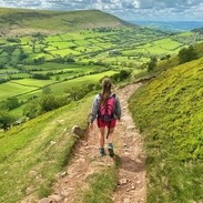 Rückenansicht einer jungen Frau, die auf einem Weg durch ein üppig grünes Tal und über Hügel geht