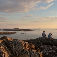 Two men sat on a rock cliff looking out to sea at dusk
