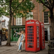 A woman leans against a red telephone box and takes a selfie in a town square.